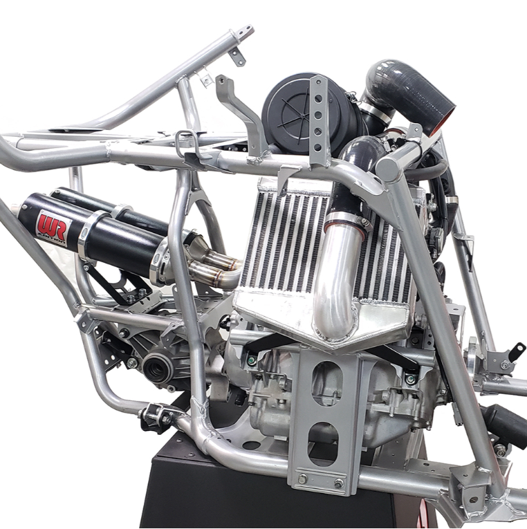 A close-up studio shot of a silver metal race car chassis and engine components mounted on a black display stand, with a white background.