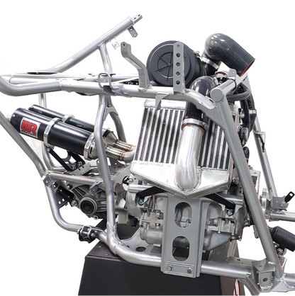A close-up studio shot of a silver metal race car chassis and engine components mounted on a black display stand, with a white background.