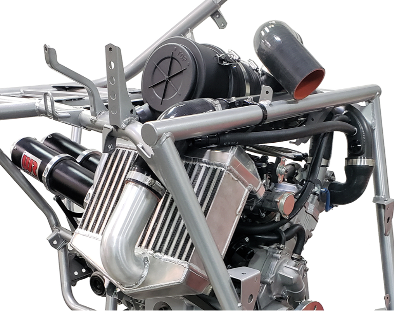 A close-up of a high-performance engine with a silver metal tubular frame, black air filter, polished silver intake manifold, and black exhaust pipes on a white background.